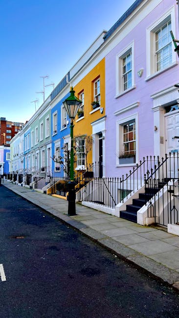 A row of colorful terraced houses in Kensington, featuring pastel facades in shades of blue, yellow, and lavender, with white-framed windows and black wrought iron railings along the front steps. The pavement is paved with concrete slabs, and there is a black street lamp positioned on the sidewalk, near one of the front doors. The houses are closely aligned, with some having small front gardens enclosed by metal fences, and others directly accessible from the sidewalk. The sky is clear with bright daylight illuminating the scene. This residential street may be part of a property relocation area, where movers like Movers Kensington could be involved in the logistics of house removals, packing, and furniture transport through narrow streets typical of London neighborhoods.