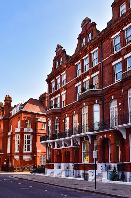 The image depicts a row of traditional red brick residential buildings with four to five floors, large sash windows, and decorative iron balconies on the upper levels. The buildings are set on a quiet street with a paved pavement, a metal street sign, and a few small plants near the entrance steps. In front of the buildings, there is a van parked on the street, partially visible, which may be used for furniture transport during a house removal. The scene is illuminated by daylight with a clear blue sky overhead. Inside one of the buildings, movers from Movers Kensington are engaged in a home relocation, visible carrying cardboard boxes wrapped in plastic, some placed on trolleys for easier loading. The loading process involves identifying furniture and boxes in preparation for transport, with staff working methodically to load items into the van, which is parked adjacent to the property. This scene reflects professional packing, moving, and furniture transport activities associated with residential removals, emphasizing careful handling and organized logistics typical of house removal services in Kensington.