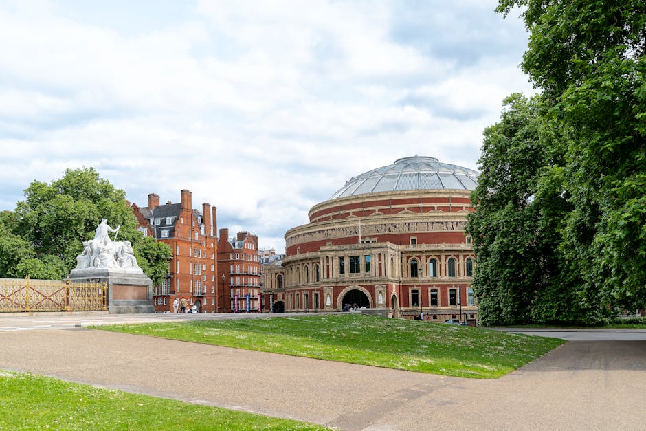 A wide outdoor view of the Royal Albert Hall in Kensington, London, capturing its circular brick and stone façade with multiple arched windows and a domed roof. In the foreground, there is a paved pathway extending across a green lawn, with a white marble statue depicting a seated figure and a sculpture of a reclining figure nearby, both on a raised platform. Surrounding the scene are tall, leafy green trees on the right and left sides, with a partly cloudy sky overhead. The setting appears to be a park area adjacent to the historic concert hall, with no people visible, emphasizing the building's architectural details and the landscaped environment, supporting house removals and relocation services nearby by illustrating the local area.