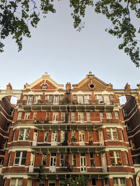 A multi-storey red brick residential building situated on a quiet city street under a clear blue sky, featuring large windows, decorative window arches, black wrought iron railings on small balcony areas, and white wooden supports beneath the overhanging sections of the upper floors. The ground floor has white steps leading to entrance doors, with a metal railing along the stairs. To the right, a black trolley loaded with cardboard boxes and wrapped furniture is visible, indicating an active home relocation or furniture transport process. The boxes are secured with plastic wrap, and the trolley is positioned on the pavement adjacent to the building. The scene captures the loading or unloading phase of house removals, with Movers Kensington likely involved in the detailed packing and moving operations. The overall environment reflects a professional and organized moving service preparing for or completing the furniture transport and packing tasks involved in a home relocation, emphasizing careful handling of household items during the loading process.