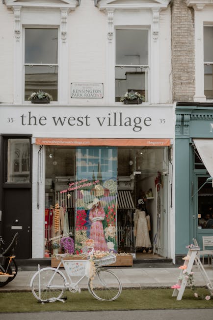 The image depicts the storefront of a shop called 'the west village' located at 35 Kensington Park Road, with large front windows displaying clothing and accessories, and an orange awning above the entrance. Outside on the pavement, there is a white bicycle with a basket parked in front of the store. Nearby, a small wooden display stand and some potted plants enhance the street scene. The upper part of the building shows windows with flower boxes underneath, with a street sign indicating 'Kensington Park Roadway.' This setting illustrates a typical street view in Kensington, relevant to local house removals and moving services. Movers Kensington, a professional removals company, may use images like this to connect their home relocation and furniture transport services with the aesthetic of the area.