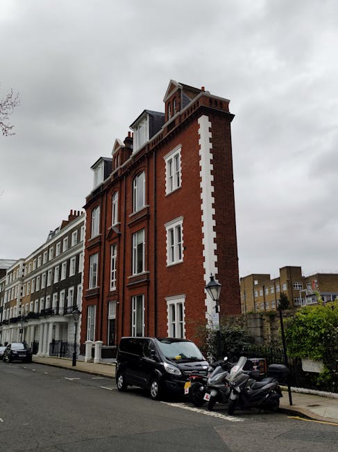 A multi-storey red brick building with white window frames and decorative architectural elements situated on a city street. The building has a pitched roof with dormer windows and is positioned next to other residential structures with similar height and classic Victorian design. In front, there is a parked black van and several motorcycles on the pavement, indicating recent or ongoing home relocation activity. The street features a lamppost and a sidewalk with some greenery, while overcast clouds cast diffused light across the scene. This setting demonstrates typical urban housing and provides context relevant to house removals and moving logistics, as managed by Movers Kensington, in line with the Kensington Church Street removal access tips for narrow homes.