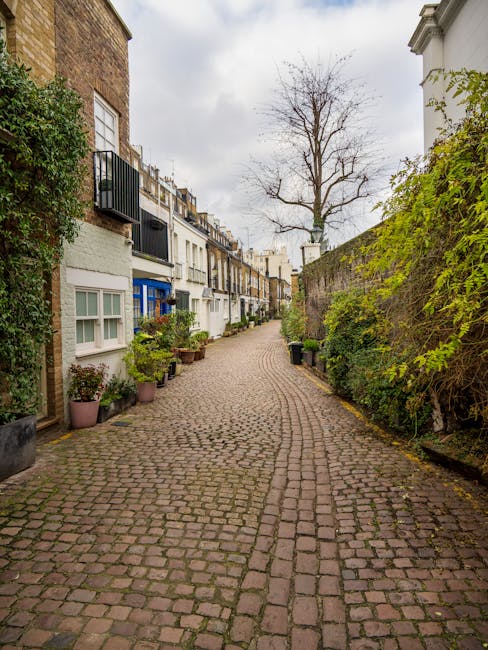 A narrow residential street with a curved cobblestone pavement, flanked by a variety of terraced houses with different facades, some with small balconies and others with large windows. Potted plants and greenery are arranged along the base of several buildings, adding a touch of nature. On the right side, a high brick and stone wall is covered with climbing plants and bushes, while on the left, the houses have white and pastel-colored exteriors with visible window frames. A leafless tree stands prominently in the background against an overcast sky, indicating a colder season. The street appears to be part of a quiet neighbourhood suitable for house removals or furniture transport, with no vehicles or people visible in the image. The scene is well-lit with natural daylight, highlighting the textures of the cobblestones and building facades. This setting exemplifies the kind of narrow residential street where professional removals companies like Movers Kensington can assist with home relocation, particularly in confined urban environments.