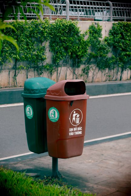 A green waste disposal bin with a black pictogram of a person disposing of rubbish, mounted on two metal legs outside a building with a yellow tiled wall. The bin is positioned on a paved area with a