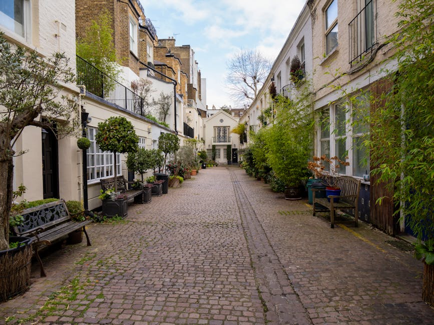 A narrow cobblestone courtyard or alleyway in a residential area, lined with white-painted terraced houses on both sides. The houses feature large windows, some with black wrought-iron balconies or window boxes filled with plants. Potted shrubs and small trees are placed along the sides, with some on black planters or in containers. Wooden benches are positioned outside some houses, and a variety of green foliage adds to the scene. The area appears to be a quiet, well-maintained outdoor space typical of London’s Kensington district. The lighting indicates daytime with natural light and partly cloudy sky. This setting may be part of a home relocation or furniture transport process, with the narrow path suitable for the loading and unloading of removal vehicles, such as those used by Movers Kensington for house removals, packing, and loading operations.