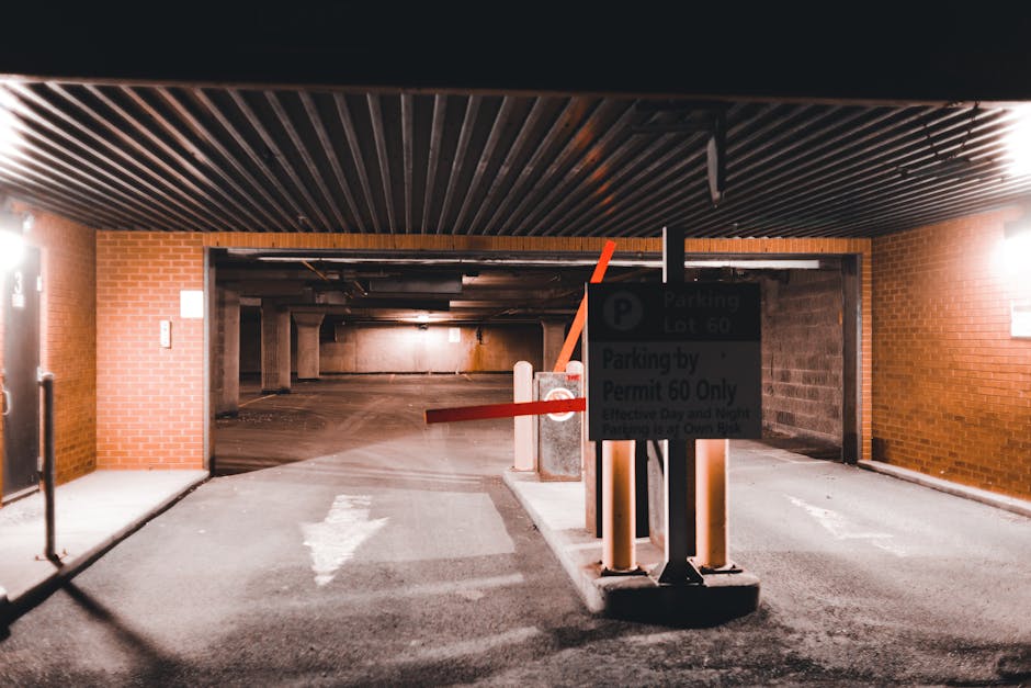 The image depicts an indoor parking garage entrance under a brick wall and a black, slatted ceiling. A white arrow painted on the concrete ground directs vehicles straight ahead towards the parking area. In front of the entrance, there is a parking permit sign mounted on a black stand, with a red barrier arm raised, indicating an active access point for authorized vehicles. The signage appears to specify restrictions related to parking permits, relevant to house removals and moving logistics managed by Movers Kensington. Inside the garage, the area is dimly lit with a warm glow, showing a spacious, concrete-floored parking lot with supporting concrete pillars and a visible vehicle pathway leading further into the building. The scene is suitable for illustrating a typical vehicle loading or unloading zone associated with residential moves, including parking regulations during a house relocation in Kensington, aligning with the service category of removals and packing and moving tasks.