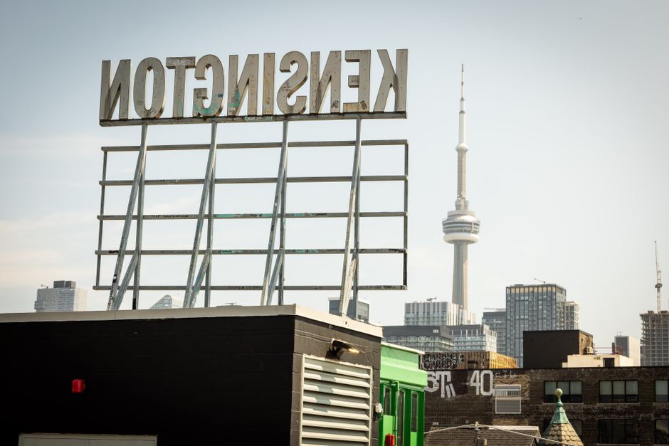 A large, weathered sign with the word 'KENSINGTON' in uppercase metal letters is mounted on a metal framework situated on the roof of a building. The sign appears to be aged, with visible rust and discoloration. The roof area underneath the sign is dark-colored, possibly black, with a small vent or louvered panel visible on the side. In the background, the London skyline features the tall, iconic BT Tower along with various modern high-rise buildings, some of which include construction cranes, indicating ongoing development. The sky is partly cloudy with soft, natural daylight illuminating the scene. Although no moving activity is visible, the setting suggests an urban environment potentially linked to property management or urban relocation services like those provided by Movers Kensington, as they handle home relocations, furniture transport, and packing during house removals in the Kensington area.