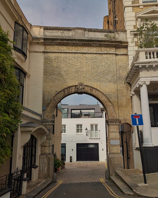 A narrow cobblestone alleyway in a residential area with brick buildings on the right side, featuring multiple white-framed sash windows and black downspouts, and a brick wall with potted plants and g